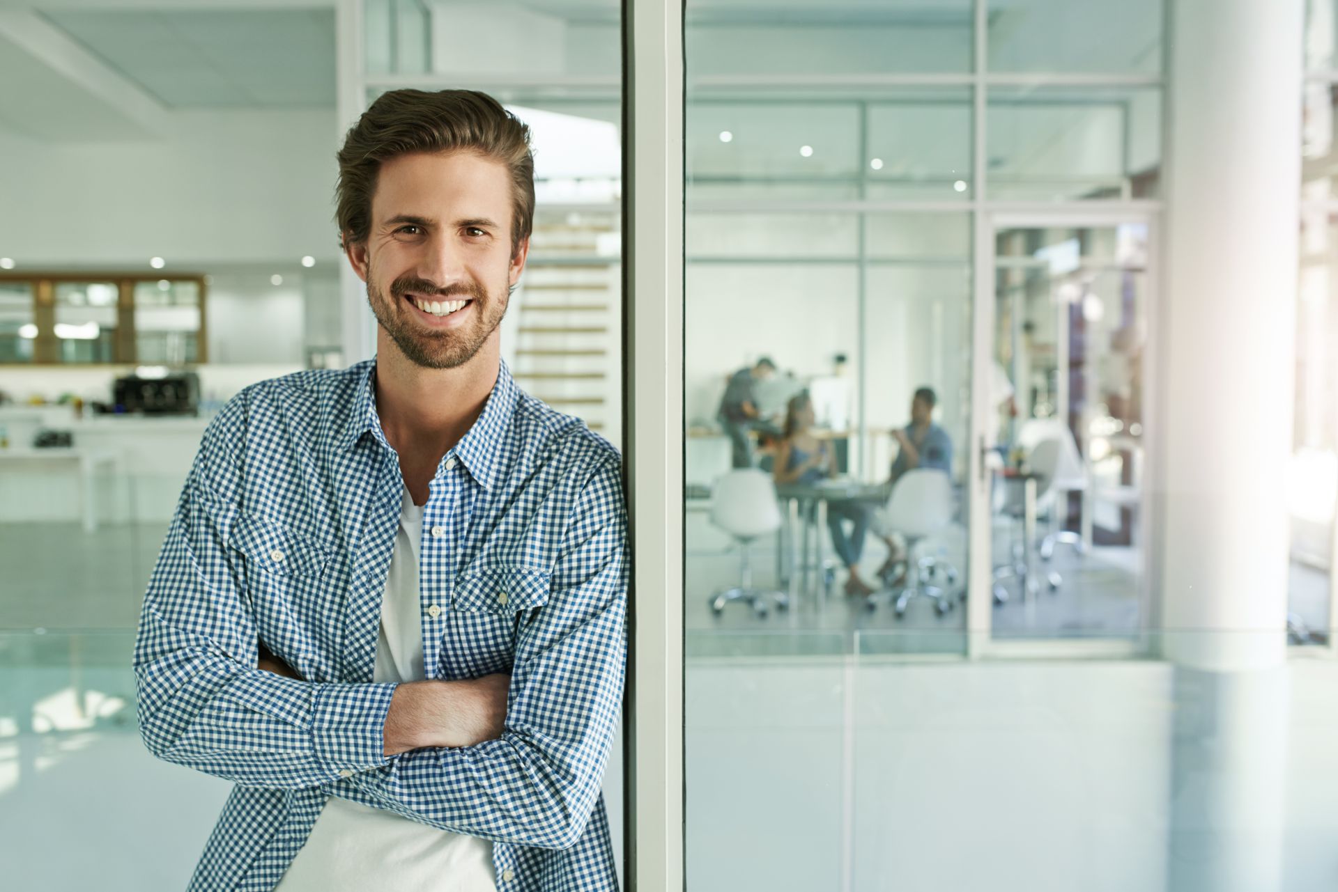 Mann im Businesslook lehnt leger an eine Trennwand im modernen Büro gelehnt und lacht in die Kamera.