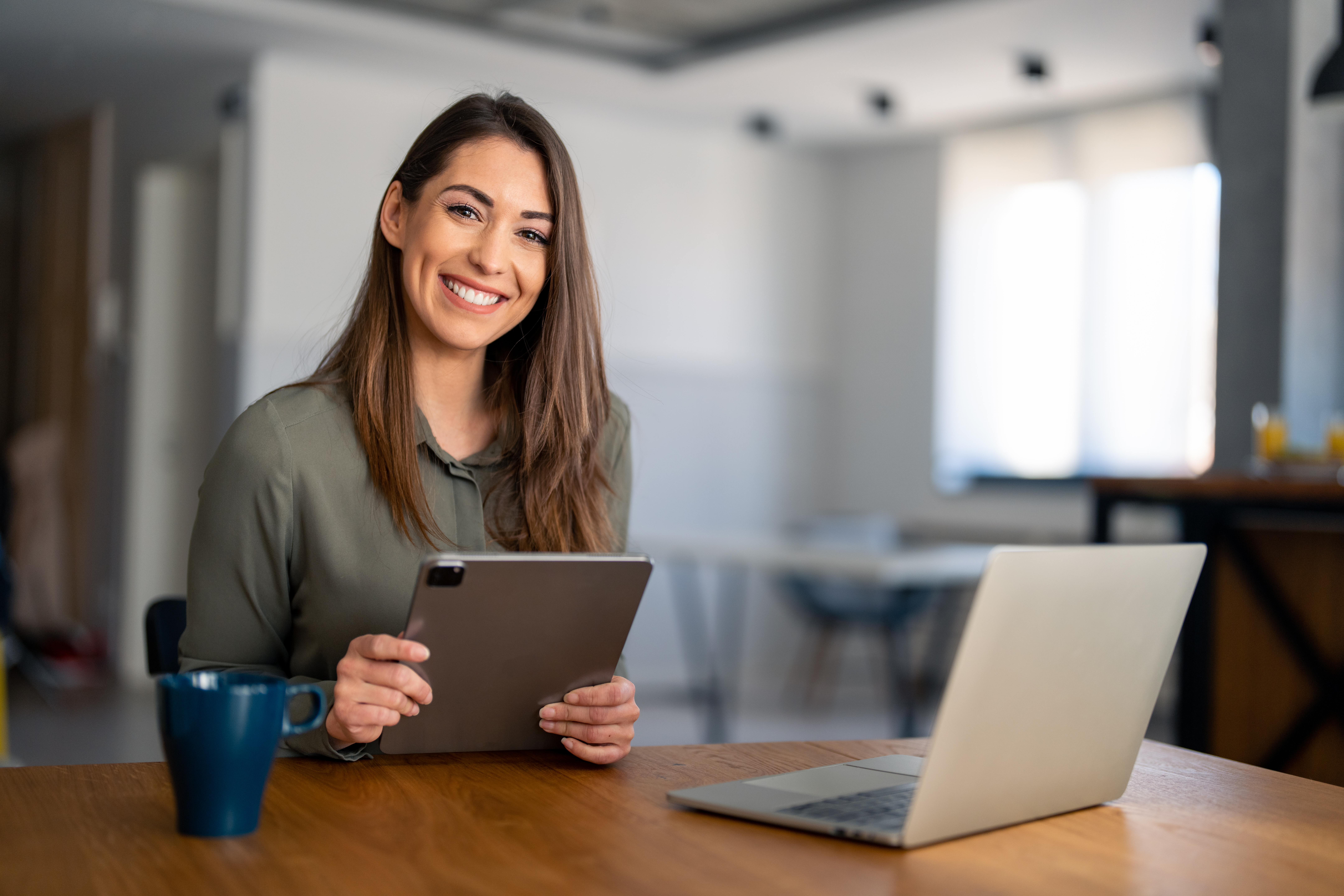 Lächelnde, selbstbewusste Geschäftsfrau, die im Homeoffice am Schreibtisch sitzt und in die Kamera blickt. Moderne, stilvolle Unternehmensmitarbeiterin und erfolgreiche Managerin, die ein Tablet hält und für ein Businessporträt posiert.